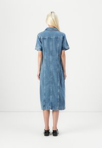 Abito camicia in denim di colore azzurro chiaro, lunghezza al ginocchio con maniche corte, chiusura frontale con bottoni e cuciture verticali. Scarpe nere completano il look.