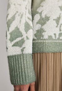 Strickpullover in Hellgrün und Creme mit Blattmustern; hat gerippte Bündchen und eine weiche Textur; getragen über einem plissee-beigen Rock.