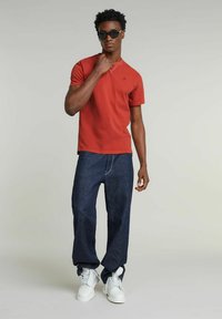 Uomo che indossa una camicia rossa a maniche corte, jeans blu scuro, stivali bianchi e occhiali da sole neri, in piedi contro uno sfondo chiaro e semplice.