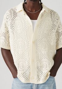 Man in een crèmekleurige, kort- mouwige opengehaakte cardigan over een witte tanktop, met een zilveren ketting, lichte jeans en handen in de zakken.