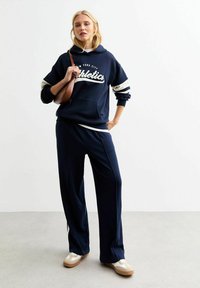 Marine hoodie met witte "Athletics" tekst, gecombineerd met bijpassende marine broek en crèmekleurige sneakers. Soepele stof en een ontspannen pasvorm.
