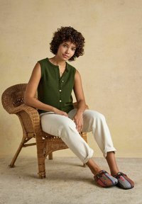 Woman with curly hair sitting in wicker chair wearing green sleeveless buttoned top, white pants, and colorful woven slip-on shoes.