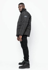 Svart puffjacka med dragkedja fram, vadderade sektioner och en logotyp-patch på vänster ärm, bärs över svarta tajta jeans och mörka stövlar.