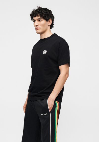 Νέος άνδρας με σγουρά μαλλιά που φοράει μαύρο t-shirt με στρογγυλό λογότυπο και μαύρη φόρμα με πράσινες, κίτρινες και κόκκινες πλευρικές ρίγες, τα χέρια στις τσέπες.