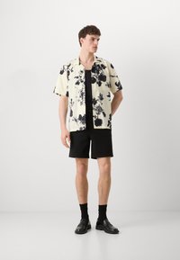 Camicia a maniche corte gialla con motivo floreale e dettagli neri, abbinata a pantaloni corti neri e scarpe nere. Tessuto morbido e vestibilità rilassata.