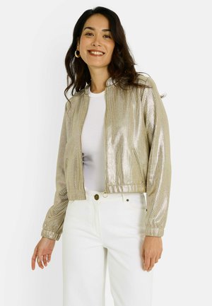 Chaqueta bomber dorada metálica con cremallera frontal, hombros estructurados y puños de punto, combinada con una camiseta blanca y pantalones de color claro.