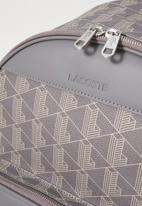 Zaino Lacoste grigio realizzato in materiale testurizzato con un motivo geometrico. Presenta doppie cerniere in metallo e un logo impresso sulla parte anteriore.
