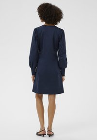 Žena s kučeravými vlasmi, oblečená v tmavomodrých dlhých šatách s nadúvanými rukávmi a čiernymi nízkymi podpätkami, stojí otočená chrbtom.