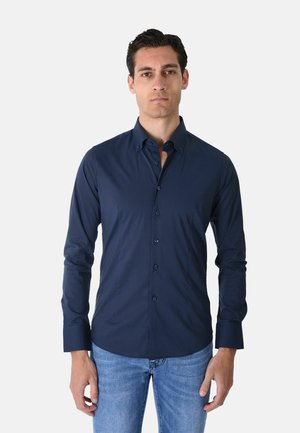 Uomo che indossa una camicia blu scuro con bottoni e maniche lunghe e jeans azzurri, in piedi davanti a uno sfondo chiaro uniforme.