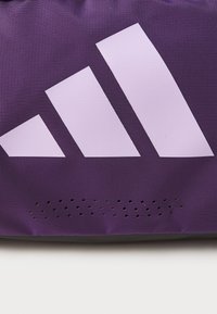 Lilla idrettsbag med teksturert overflate, utstyrt med en stor hvit tre-stripe logo og små hull nær bunnen.