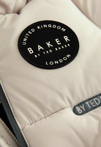 Beige gepolsterte Jacke mit einem runden schwarzen Stoffaufnäher. Der Aufnäher zeigt weißen Text: "BAKER by TED BAKER," mit "VEREINIGTES KÖNIGREICH" und "LONDON."