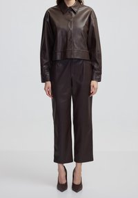 Giacca cropped in pelle marrone con chiusura a bottoni e colletto intrecciato, abbinata a pantaloni ampi marroni e décolleté a punta.