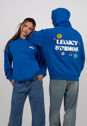 Blue hoodies made of soft fabric, featuring 'Legacy Studios' in white on the back and multicolored floral designs. Front has small logo.