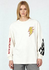 Femme blonde portant un chemisier blanc à manches longues avec un éclair jaune sur la poitrine, le texte rouge « VOUS AVEZ LE POUVOIR » sur la manche gauche, et une flamme noire sur la manche droite.