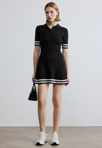 Melna ribota polo kleita ar īsām piedurknēm, baltas svītras uz piedurknēm un apakšmalē, kombinēta ar baltām sporta kurpēm un mazu melnu somiņu.