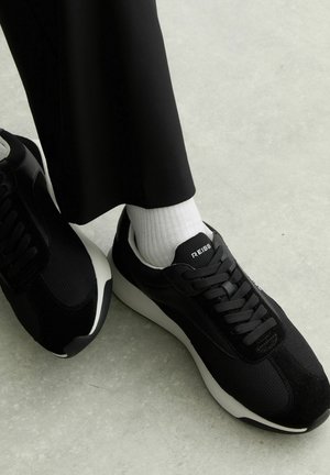 Fekete Reiss tornacipők fehér talppal, fehér zoknival és fekete nadrággal, világosszürke padlón állva.