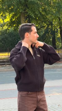 Pulover tricotat, de culoare maro închis, cu fermoar și guler înalt, având o textură ribbed, asortat cu pantaloni maro deschis, în exterior, într-un mediu verde.