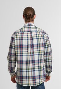 Mann mit nach hinten gebundenem Haar, der ein mehrfarbiges kariertes Langarmhemd und blaue Jeans trägt, mit dem Gesicht von einer einfarbigen Hintergrund abgewandt.