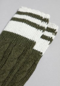 Olivenfarvede fingerløse handsker i ribstrik med hvide vandrette striber ved håndleddene. Blød tekstur med synlige syninger.