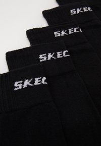 Melni zeķes ar ribotām malām, uz kurām ir balts adīts "SKECH" teksts joslā. Mīksta tekstūra un vienkāršs dizains.