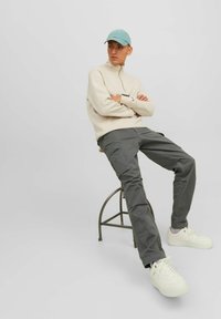 Hellbeige Zip-Up-Pullover, graue Cargo-Hosen und weiße Sneaker. Das Model trägt eine hellblaue Mütze und sitzt auf einem Metallhocker.