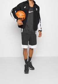 Person in Nike sportswear holding an orange basketball, wearing black and white jacket, shorts, black socks, and black sneakers, standing indoors.
