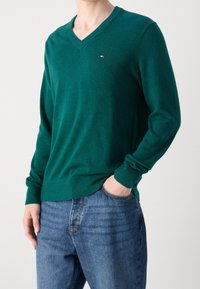 Maglione verde a scollo a V realizzato in materiale morbido, con maniche lunghe e polsini a coste, indossato sopra jeans blu. Piccolo dettaglio del logo sul petto.