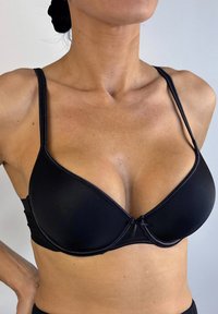 Sostén negro con aros, que presenta una tela suave, tirantes ajustables, una forma redondeada y un pequeño lazo decorativo en el centro frontal.