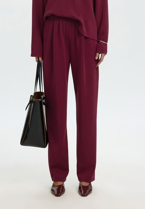 Femme portant un pantalon bordeaux ample et un haut assorti, tenant un grand sac noir et portant des chaussures bordeaux à bout pointu.