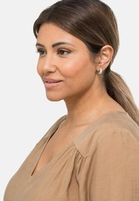 Pendientes de plata con acentos similares a cristales, sobre un tono de piel suave y claro. El modelo lleva una blusa beige de diseño con escote en V.