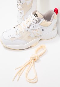 Baskets blanches et beiges avec une tige texturée, présentant un logo "N" proéminent, des accents gris et un onglet de talon rouge. Comprend des lacets en beige clair noués.