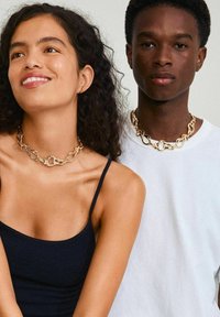 Collares de cadena en tono dorado con eslabones entrelazados; uno presenta un detalle de cerradura. La modelo lleva una blusa negra, y otra viste una camisa blanca.