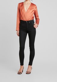 Rust satäng långärmad inlindad bodysuit i kombination med högmidjade svarta skinny jeans. Modellen bär svarta klackar med öppen tå. Slät textur.