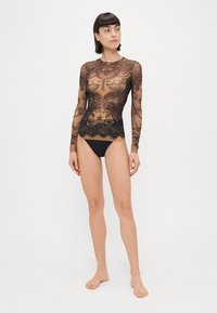 Crna čipkasta bluzica dugih rukava s složenim uzorcima i valovitim rubom, uparedita s jednostavnim crnim badi bottomom.