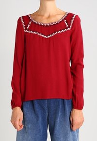 Rode blouse met lange mouwen, featuring een ronde halslijn met decoratieve borduursels en elastische manchetten, gecombineerd met losse blauwe spijkerbroek.