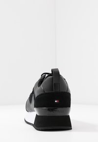 Baskets noires avec un mélange de cuir et de daim, une semelle blanche, des accents dorés et un logo sur le talon avec un détail en blocs de couleur.