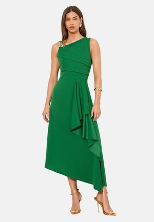 Frau trägt ein ärmelloses, asymmetrisches grünes Kleid mit einer seitlichen Rüsche, eine goldene Brosche an der Schulter, ein goldenes Armband, Ohrringe und Absatzschuhe.