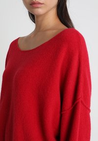 Röd överdimensionerad tröja med vid halsringning, tillverkad av ett mjukt, texturerat tyg. Droppaxel-design och långa ärmar.