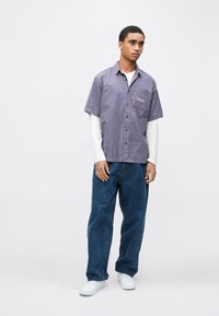 Rövid ujjú, gombos világos lila pamut ing mellzsebbel és hímzett részlettel, laza kék farmernadrággal és fehér cipővel párosítva.