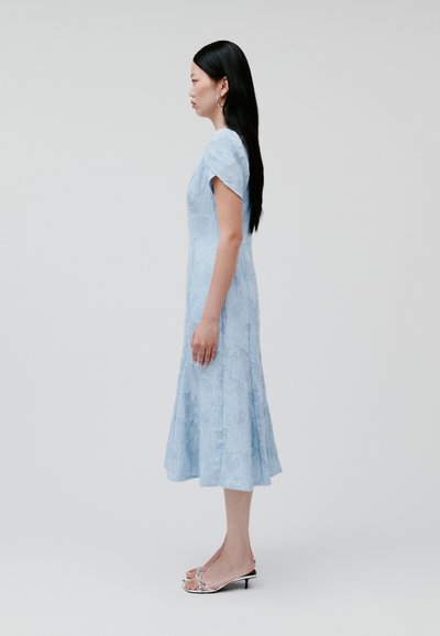 Himmelblaues Spitzekleid mit kurzen Ärmeln, tailliertem Oberteil, ausgestelltem Saum und detailliertem Strukturmuster. Getragen mit silbernen Absatzsandalen.