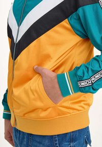Retro zip-up jacka med en färgblockdesign i orange, teal, svart och vitt. Inkluderar fickor och ribbade ärmslut med varumärkesdetaljer.