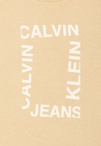 T-shirt in cotone beige con testo bianco in grassetto disposto in un motivo quadrato, con la scritta "CALVIN KLEIN JEANS" in evidenza.