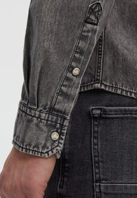 Grau denim Jackenärmel mit einem Knopfverschluss, ausgestattet mit einem strukturierten Stoff und genähten Akzenten. Enthält eine sichtbare Tasche auf einem dunkleren Hintergrund.