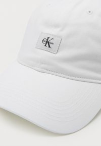 Cappello da baseball bianco in cotone con visiera curva. Presenta un'etichetta rettangolare con la scritta "Calvin Klein" e un accento del logo nero.