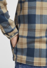 Flanellhemd mit einem blau-beigen Karomuster, aus weichem Stoff gefertigt, mit Knopfmanschetten und einer Seitentasche.