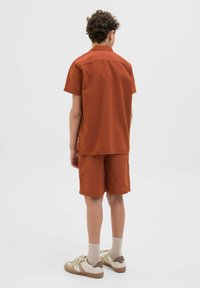 Korte mouw rustkleurig shirt en bijpassende korte broek van textuurstof. Draagt beige sneakers en lichte sokken, gezien vanuit de achterkant.