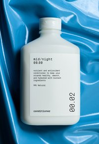 Hvit plastflaske med skrukork, med svart tekst på en etikett. Inneholder balsam, beskrevet som 98 % naturlig med bioteknologiske ingredienser.