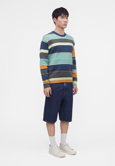 Jeune homme debout portant un pull rayé multicolore, un short en jean foncé longueur genou, des chaussettes blanches et des baskets blanches sur fond blanc.