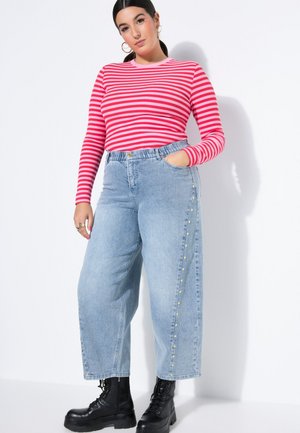 Donna che indossa un top a maniche lunghe a righe rosa e rosse, jeans azzurri a gamba larga con borchie laterali e stivali neri stringati, in piedi contro un muro bianco.