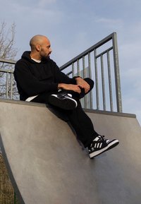 Man in black hoodie and pants sitting on the edge of a concrete skate ramp, looking to his left under a clear sky.